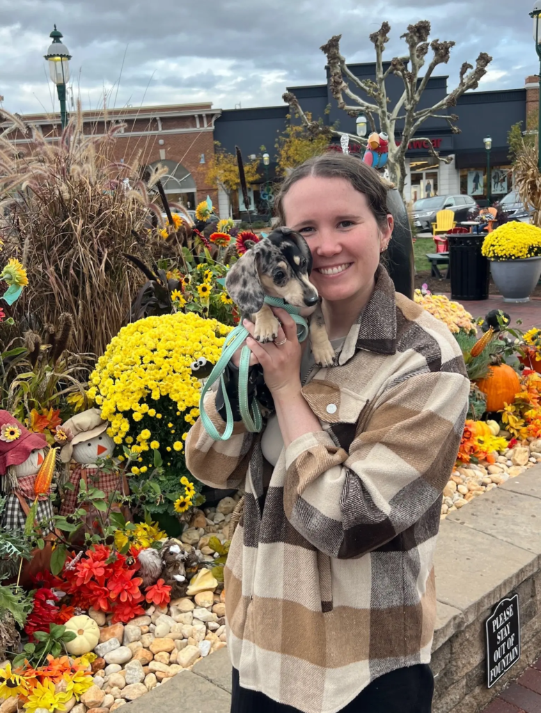 Olivia, weight-inclusive website designer, posing in front of fall flowers with her dachshund puppy